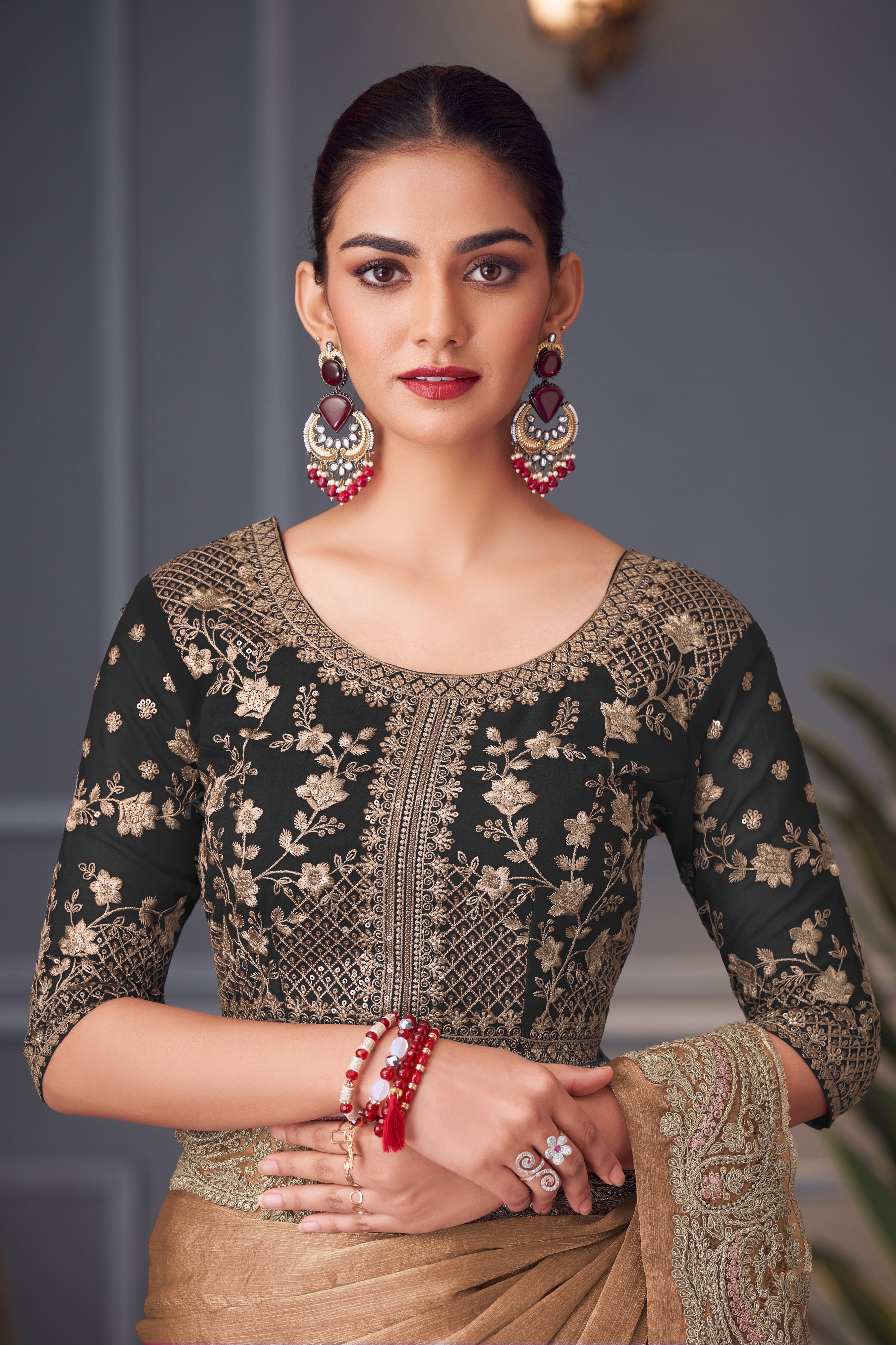 Woman wearing a black and gold embroidered blouse with a beige saree, sitting against a neutral background.